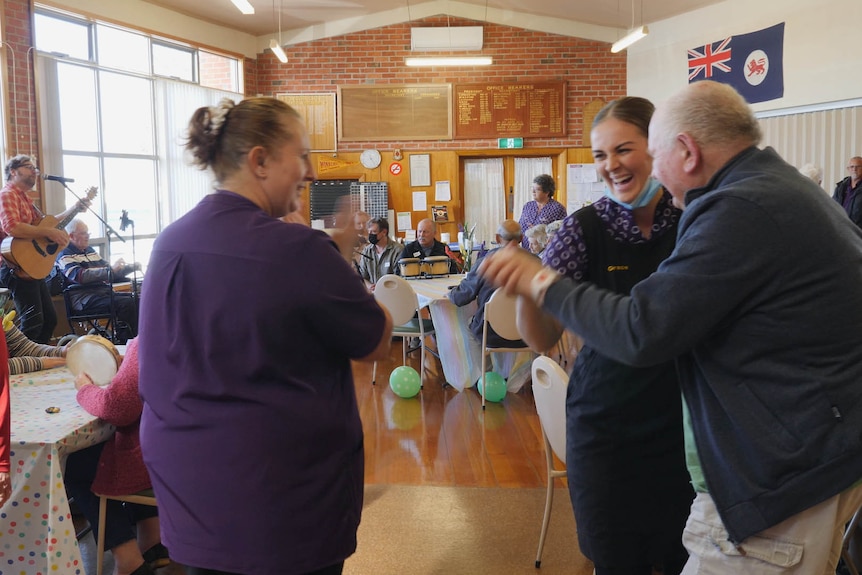 A young aged care staffer dances with elderly man to live guitarist in an indoor space