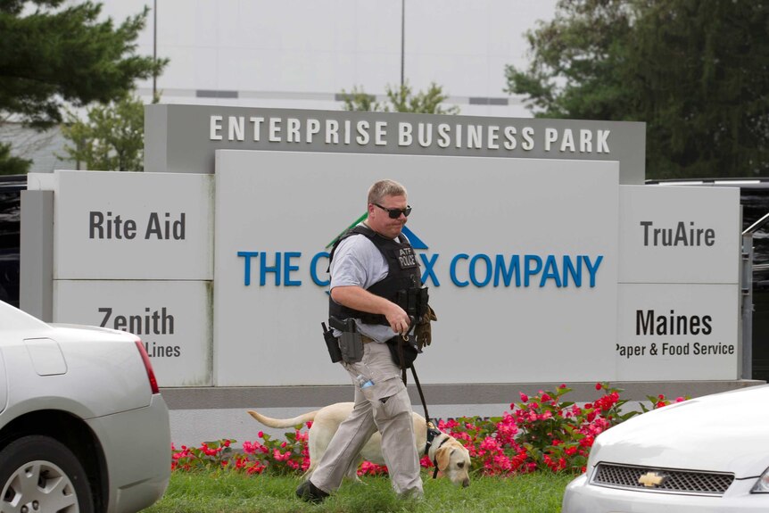 A greying man with a black vest and a holster walks a dog past a large business park sign