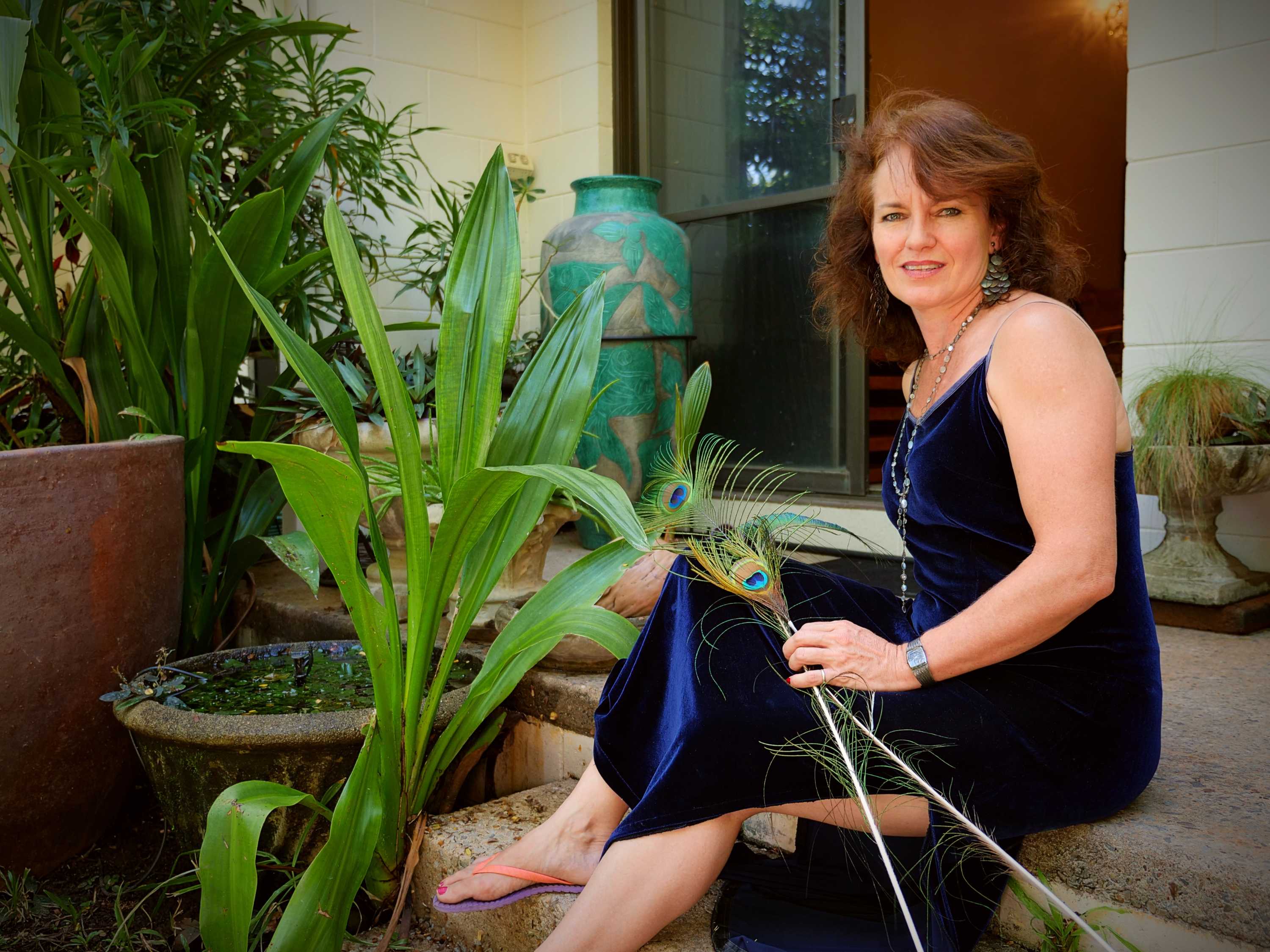 woman looing at camera wearing blue gown sitting on the back step of her house holding peacock feathers
