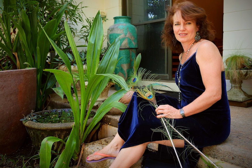 woman looing at camera wearing blue gown sitting on the back step of her house holding peacock feathers