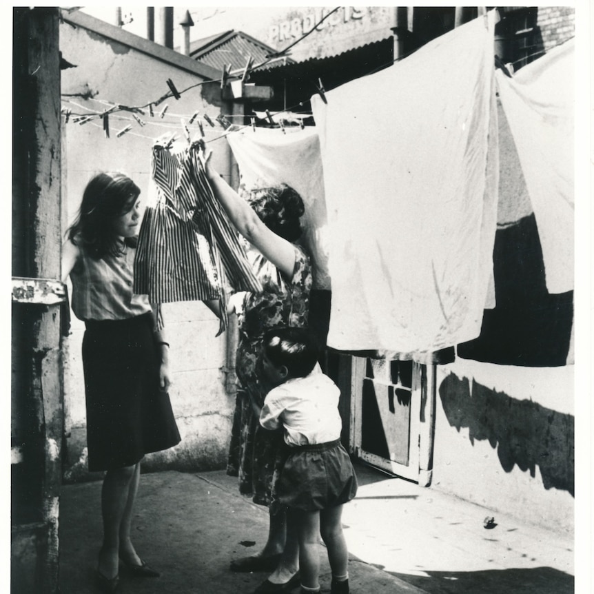 black and white photo of a woman standing next to another woman hanging clothes on the line with small child at her leg