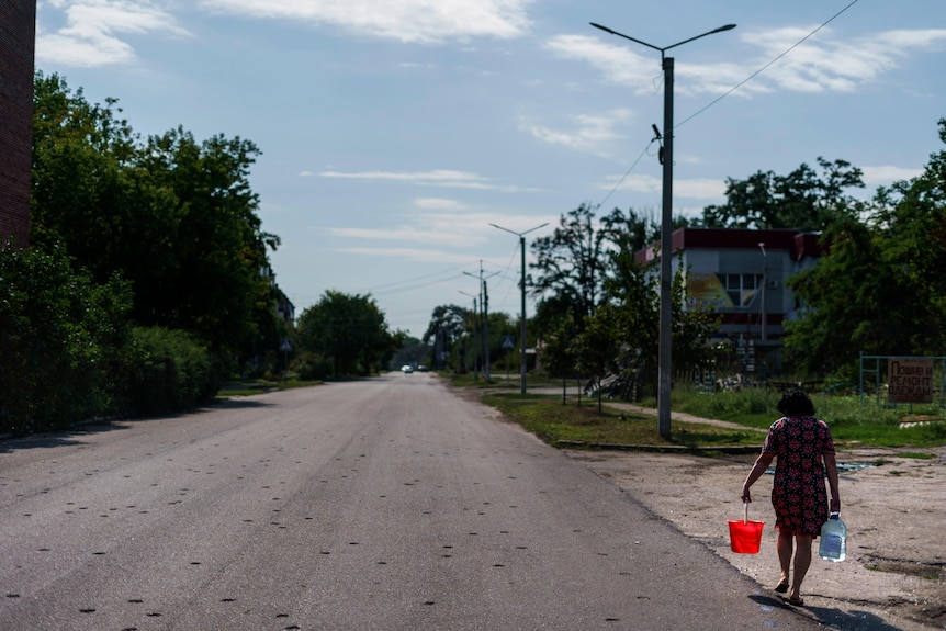 A woman walks down a road carrying two buckets. The road is spotted with damage 
