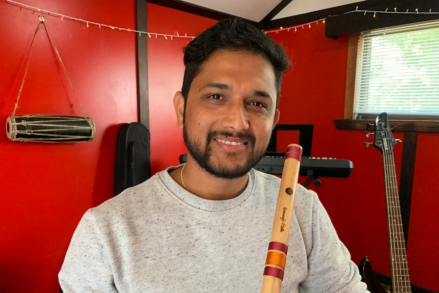 Pratik Sigdel holds a woodwind instrument as he smiles at the camera in front of a red wall.