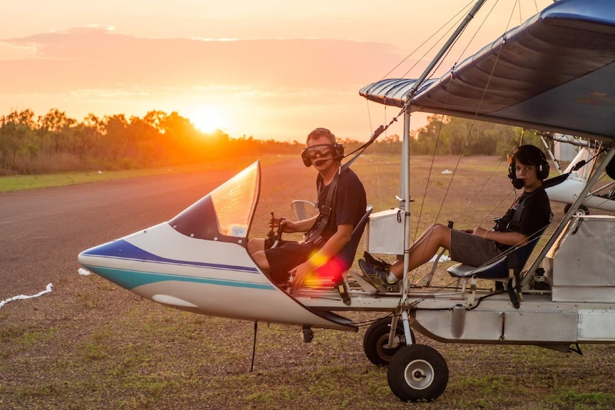 Two young boys sit in a small blue and white opencockpit aircraft with headsets on as the sun sets behind them.