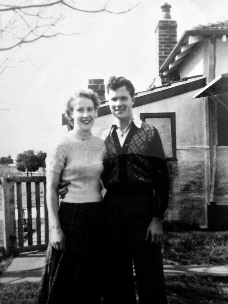 Black and white photo of a young woman and man standing in a house's front yard, their arms around each other.
