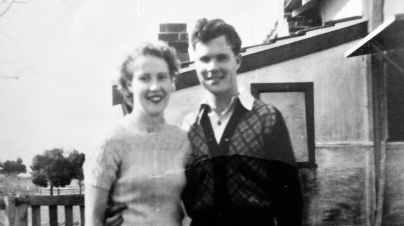 Black and white photo of a young woman and man standing in a house's front yard, their arms around each other.