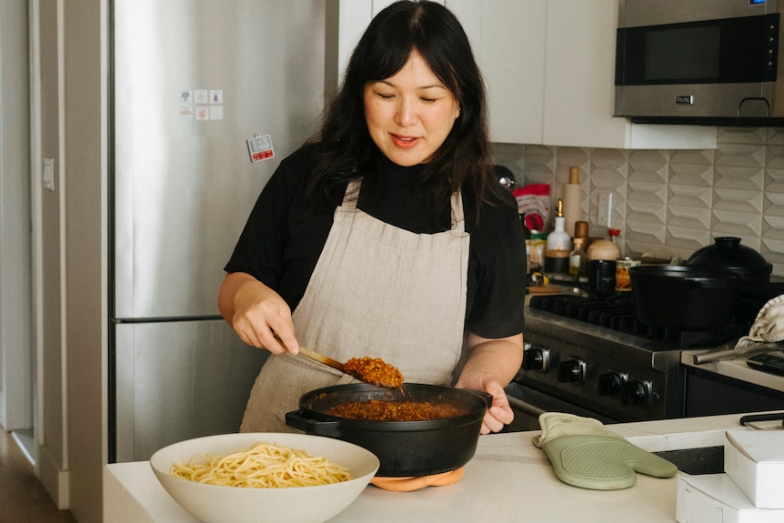 Hetty Lui McKinnon in her kitchen serving lentil bolognese ragu from a pot into a large bowl containing cooked spagetti.