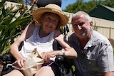 A woman wearing a sunhat sits in a wheelchair in a backyard garden, a man in a button up shirt is kneeling next to her