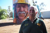 White male with big moustache stands in front of brightly painted water tower, with artist painting on lifter in background.