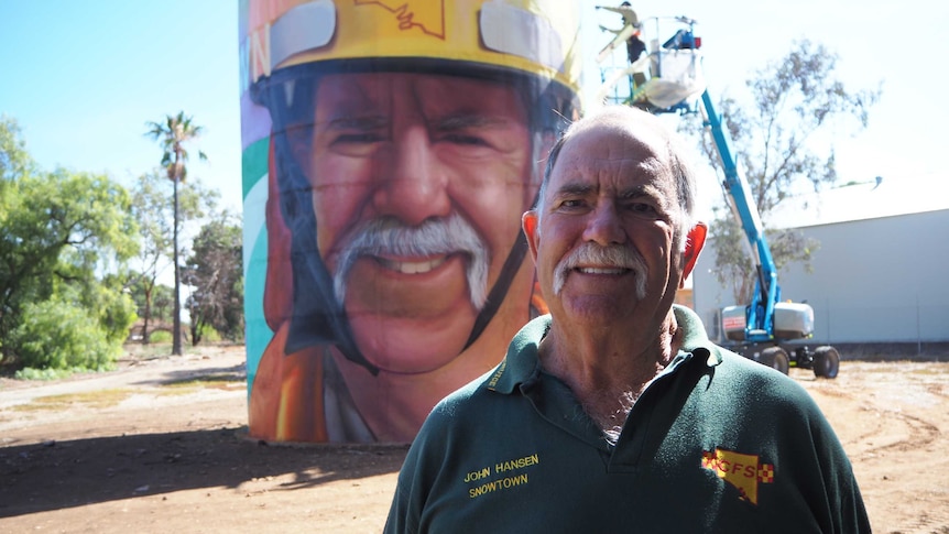 White male with big moustache stands in front of brightly painted water tower, with artist painting on lifter in background.