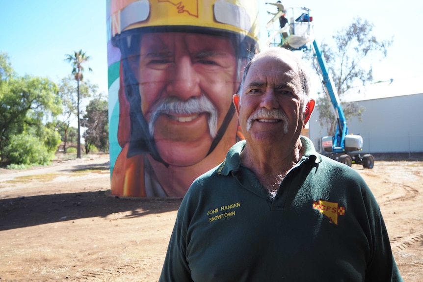 White male with big moustache stands in front of brightly painted water tower, with artist painting on lifter in background.