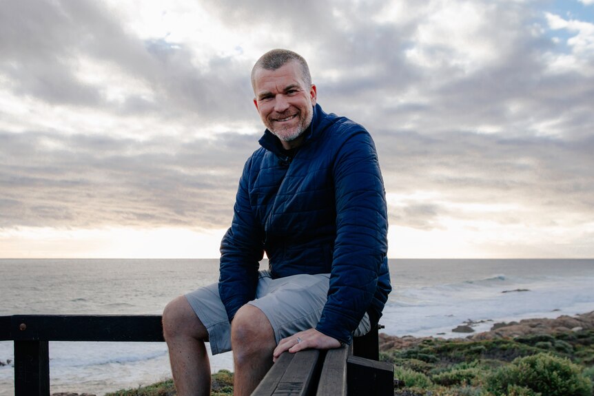 A smiling man with short grey hair sits in front of the ocean.