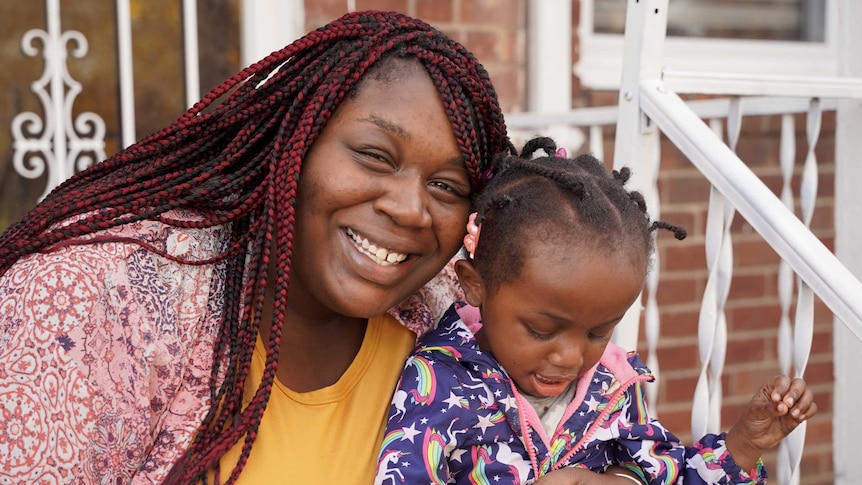 A black woman with brown and red dreads smiles with her daughter on some stairs
