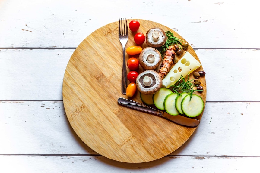 A round chopping board with food grouped in a third of the area, with a knife and fork as "clock hands".