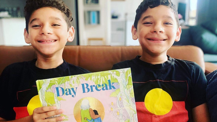 Twins Vernon and George Bond holding the children's book Day Break, smiling, wearing Aboriginal flag t-shirts.