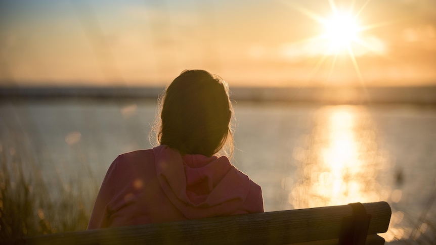 A woman sits on a park bench, her back to the camera, as the sun sets over water
