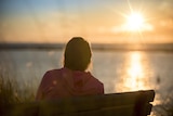 A woman sits on a park bench, her back to the camera, as the sun sets over water