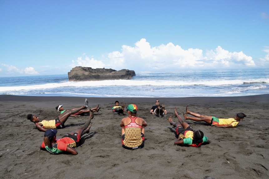 Women lie on sand in circle lifting legs, with small island in the water in the distance.