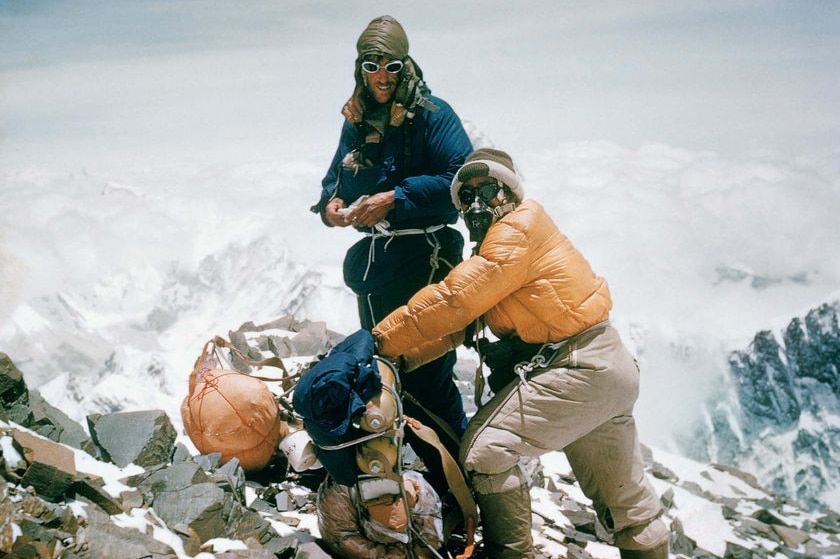 Two men stand on a tall snowy mountain. 