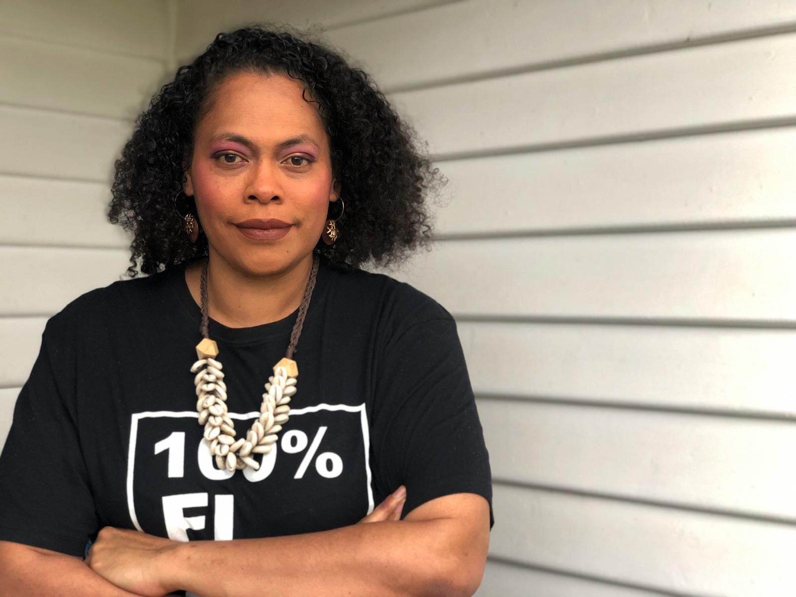 A Fijian woman in a t-shirt folds arms and looks at the camera. 