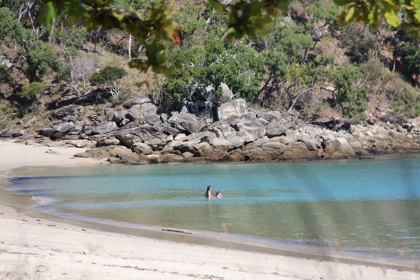 Two children swimming in the ocean off in the distance in blue waters, surrounded by white sand.