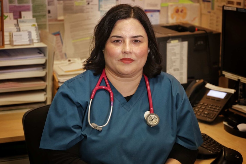 Dr Maria Boulton wearing blue-green medical shirt and stethoscope sitting at a desk in her consulting room