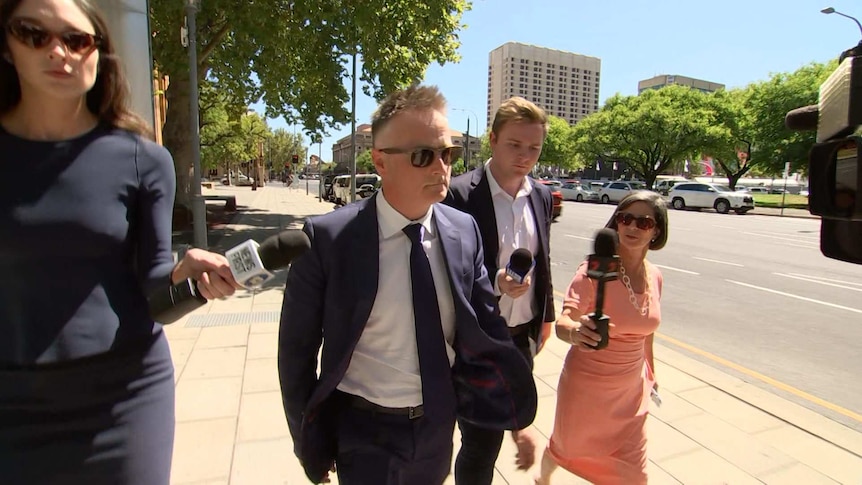 A man in a blue suit and glasses surrounded by reporters walking quickly