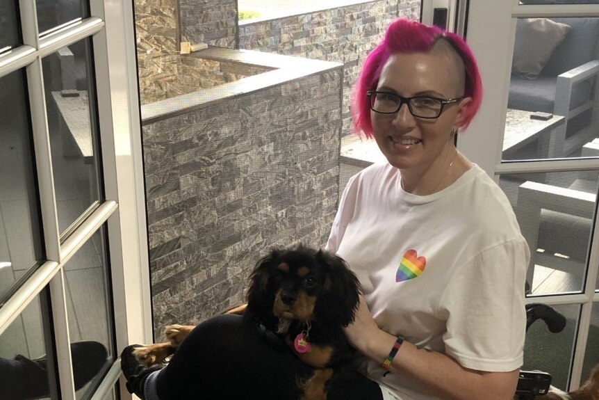 A woman with pink hair in a wheelchair holding a dog. 