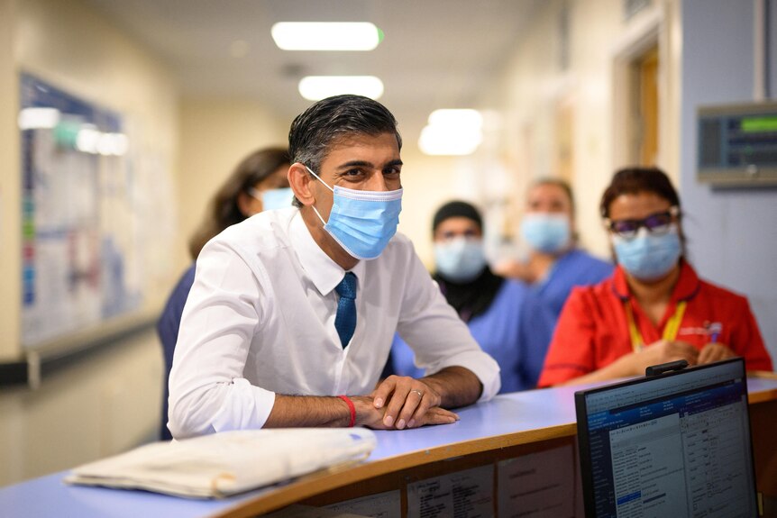 Rishi Sunak in a face mask leans on a counter at a nurses' station