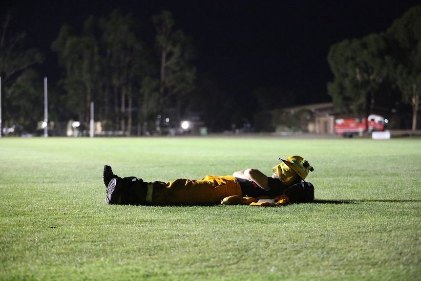 A man wearing firefighting gear uses his helmet to cover his eyes as he lies on the grass of an oval.