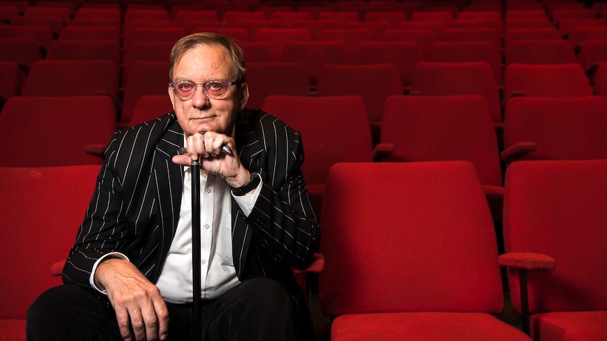 A man wearing glasses and a black striped jacket, sitting in the front row of a theatre with red seats.