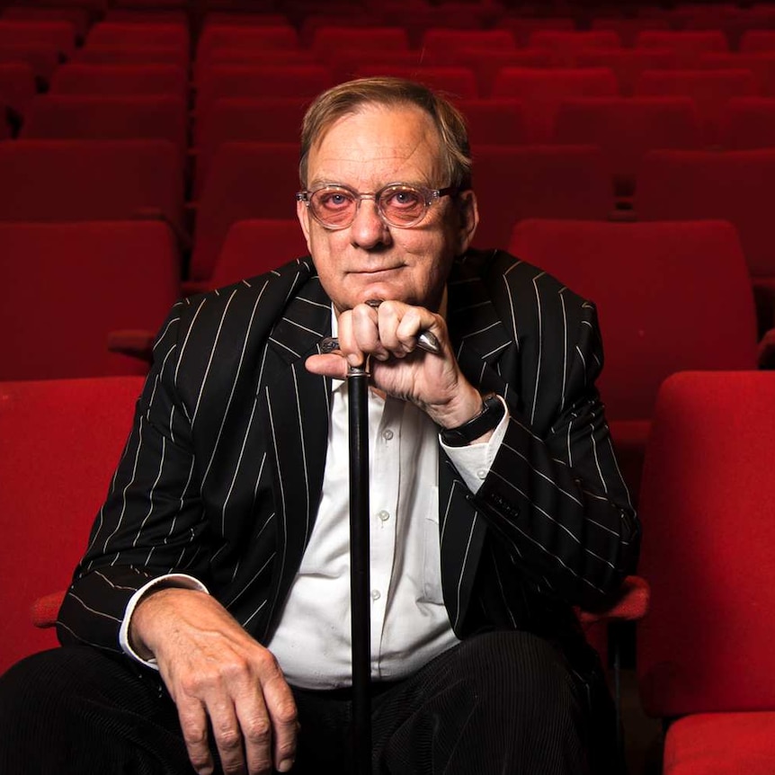 A man wearing glasses and a black striped jacket, sitting in the front row of a theatre with red seats.