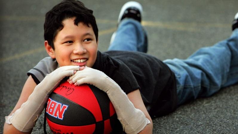 A young, smiling Terry Vo with arms bandaged, lies on a basketball court holding a basketball.