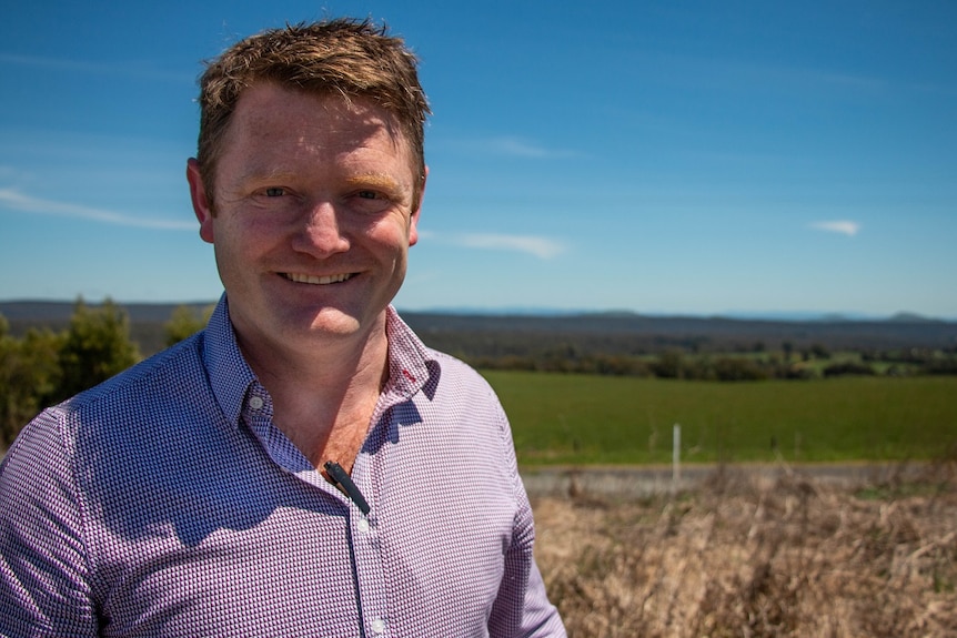 a man with short red hair stands in front of a green paddock