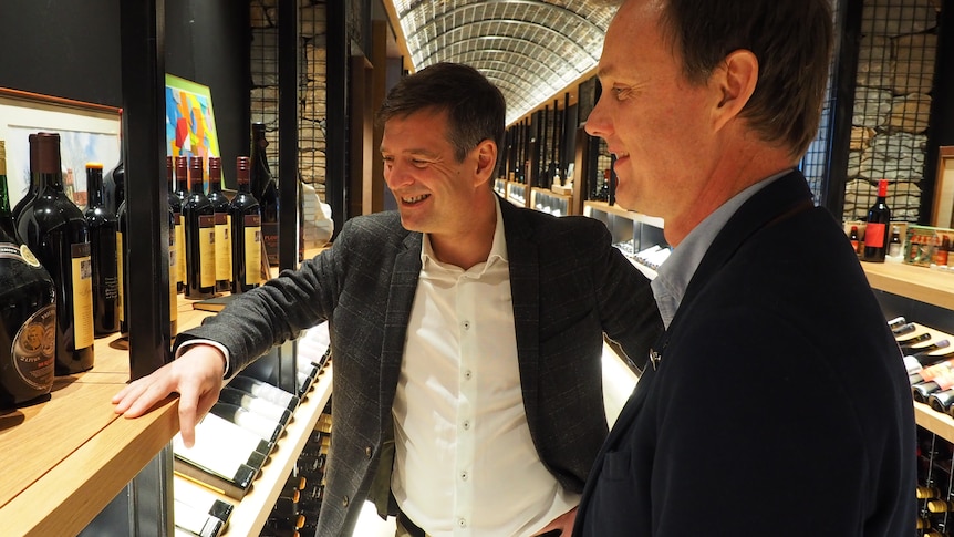 two men stand in a winery smiling and looking at bottles on a shelf