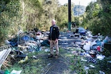 A man standing among piles of rubbish in the bush.