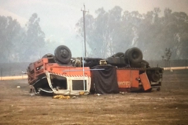 An upside down fire truck lies in a burnt paddock, with the roof smashed in.