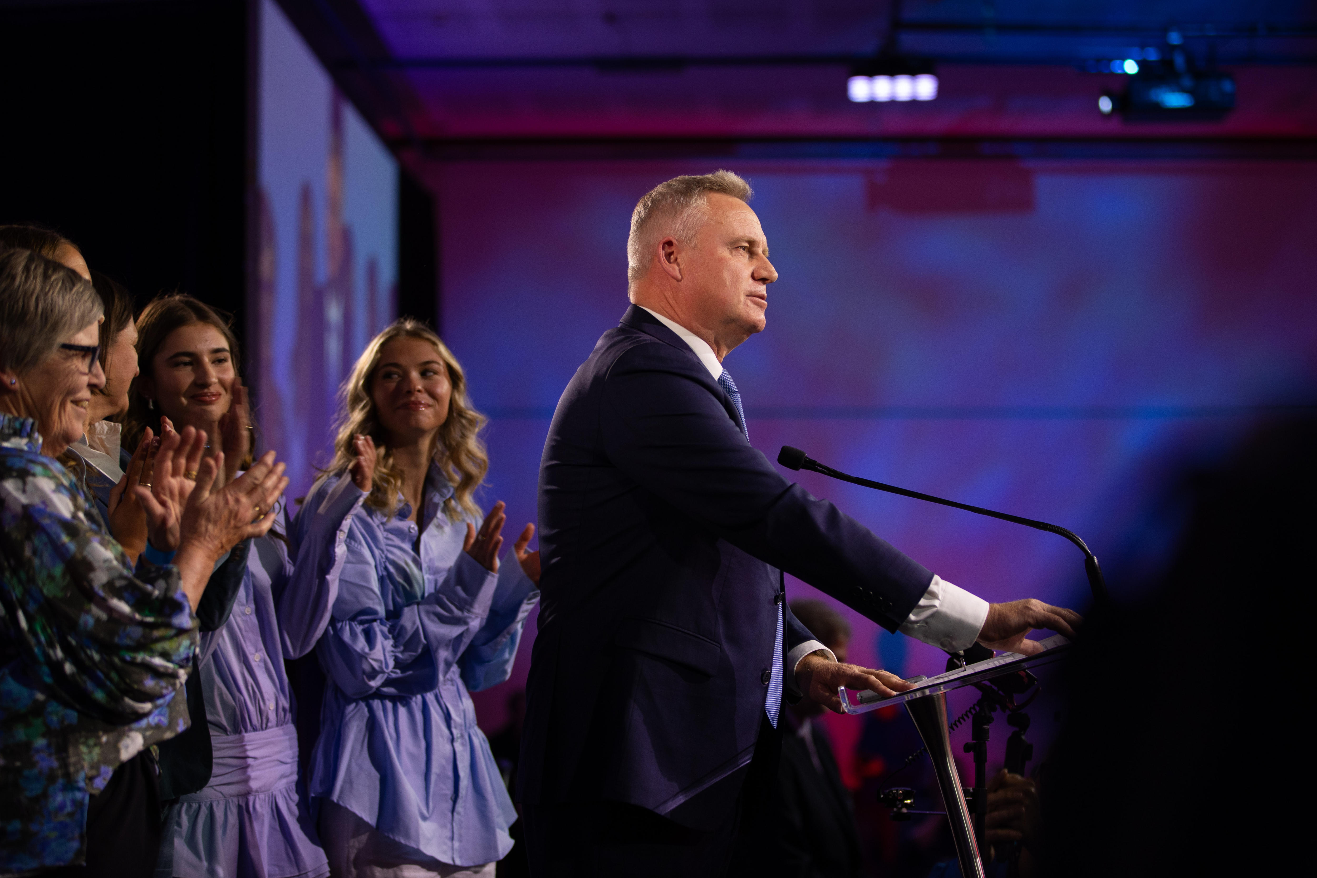 Caucasian man in suit with short hair stands at podium making speech with family behind him, looks optimistic