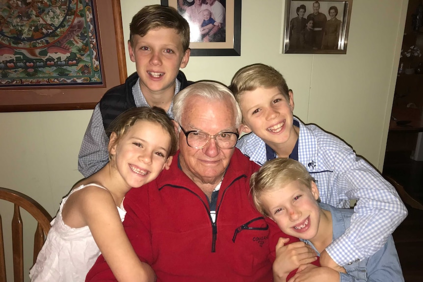 An elderly man sitting at a table with four of his grandchildren hugging him.
