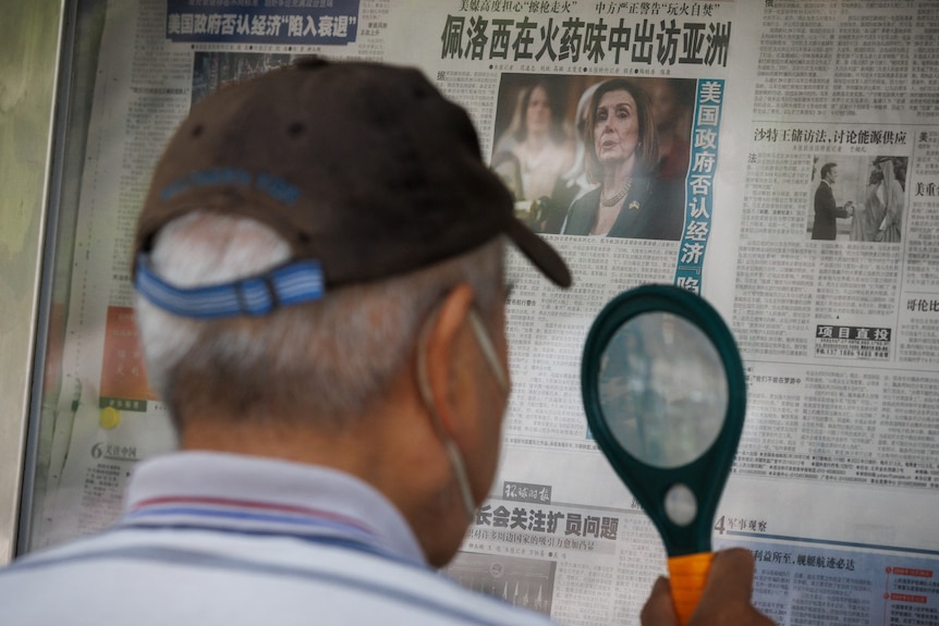 The back view of a man with grey hair wearing a dark cap, looking through a magnifying glass reading a newspaper on a stand