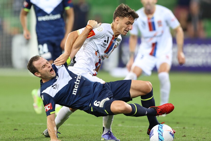 Two soccer players, one in dark blue and one in white, challenge for the ball as one pushes the other over in the grass