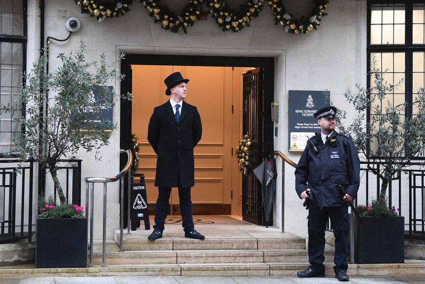 A man in a black top hat and a police officer stand outside a hospital entrance, with Christmas decorations above the door.