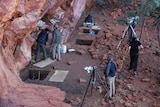 People examine rock art on an overhang while others examine a square archeological dig out. 