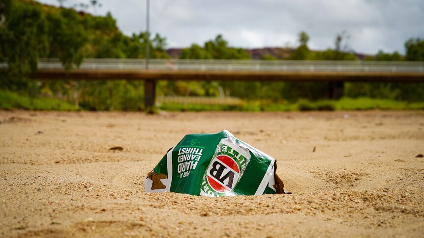An empty VB carton lying on the sandy ground, with a bridge in the distance.