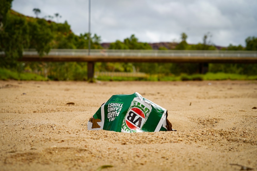 An empty VB carton lying on the sandy ground, with a bridge in the distance.