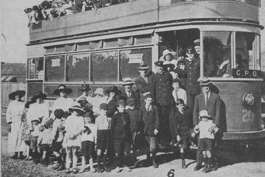 A crowd of people stand on and around a tram in this old photo.