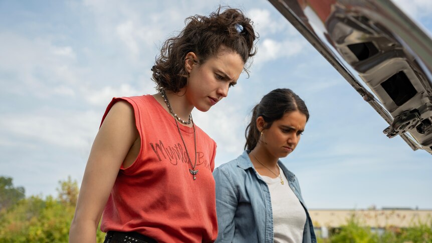 A film still of Margaret Qualley and Geraldine Viswanathan. They're looking down into the boot of a car, with confused looks.