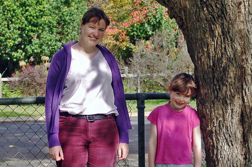 A woman and a child stand in front and a fence, next to a tree.