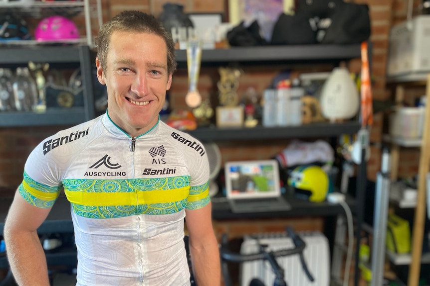 A man in cycling gear stands smiling in a garage. 