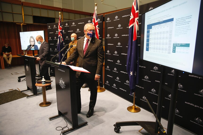The Prime Minister, wearing an Australian flag mask, stands at a lectern with a powerpoint presentation beside him.